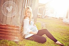 a pregnant woman sitting on the ground next to a barn with her hands in her pockets