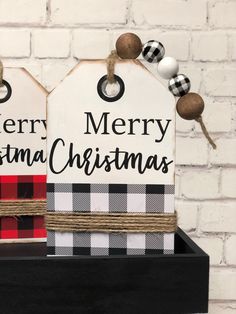 two christmas tags are sitting on top of a black shelf next to a brick wall