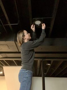 a woman in grey shirt and blue jeans standing on a black ceiling with a remote control