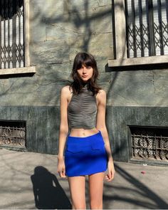 a woman standing in front of a building wearing a blue skirt and grey top with her hands on her hips