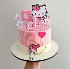 a hello kitty birthday cake with pink icing and hearts on top, is being held up by a woman's hand