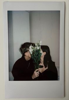 two people kissing each other with flowers in front of their face and behind them is a white wall