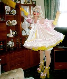 a woman dressed as a drag queen posing for the camera in her living room with lots of clutter on the shelves