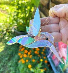 a hand holding a colorful bird ornament in it's left hand, with orange flowers in the background