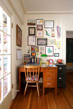 a wooden desk sitting under a window next to a wall filled with pictures and books