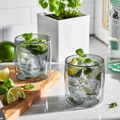 two glasses filled with ice and limes sitting on a counter next to a cutting board