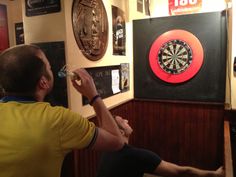 a man sitting at a table drinking from a glass in front of darts on the wall