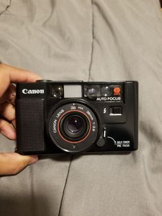 a person holding a camera in their hand on top of a bed with a gray sheet