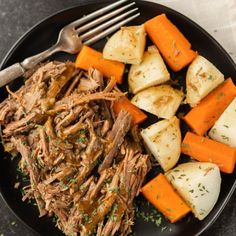 a black plate topped with meat, potatoes and carrots next to a silver fork