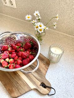 strawberries in a colander on a cutting board next to a vase with daisies