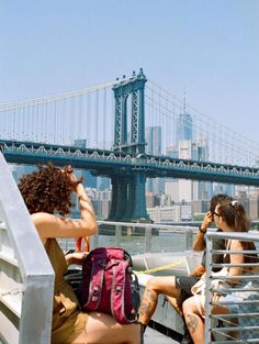 two women sitting on the side of a boat looking out at the water with a bridge in the background