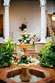 a fountain with flowers on it in the middle of a courtyard surrounded by greenery