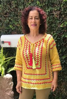 a woman standing in front of a bush wearing a yellow crochet top with red tassels