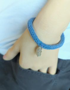 This hand-knitted blue bracelet is adorned with a Pecos Diamond quartz crystal charm. The bracelet is crafted from a soft, blue yarn, giving it a cozy and handmade appearance. The charm, which is a Pecos Diamond quartz crystal, is known for its metaphysical properties, including healing, meditation, and protection. The crystal is wrapped in a wire, enhancing its aesthetic appeal and securing it to the bracelet. This unique piece combines the comforting texture of knitted fabric with the spiritual benefits of a healing stone, making it a meaningful accessory for those interested in metaphysical practices. The bracelet is worn on the wrist, with the charm hanging delicately, adding a touch of elegance and significance to the overall design. Knitted Bracelet, Metaphysical Gifts, Metaphysical Jewelry, Knit Bracelet, Witch Gift, Protection Amulet, Diamond Quartz, Healing Meditation, Crystal Charm