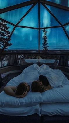 two people laying on a bed in a room with a glass ceiling and aurora lights