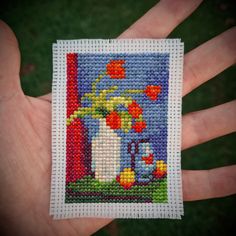 a hand is holding a small cross stitch picture with flowers in the vase and watering can