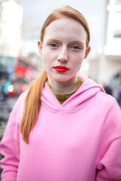 a woman with red hair wearing a pink hoodie and posing for the camera on a city street