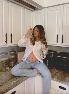 a pregnant woman sitting on top of a kitchen counter holding a cup in her right hand