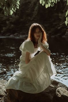 a woman sitting on top of a rock next to a river wearing a white dress