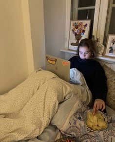 a woman sitting on a bed with a laptop and chips in front of her,