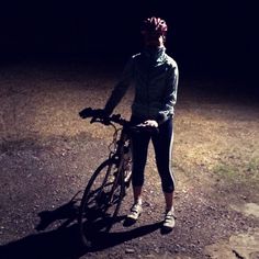 a woman is standing next to her bike in the dark, with one hand on the handlebars