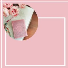 a pink book sitting on top of a wooden table next to flowers and a white frame