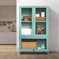 a kitchen with white walls and wooden floors, including a teal bookcase that has baskets on it