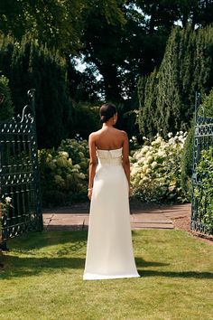 a woman in a white dress is standing on the grass near an iron gate and flowers