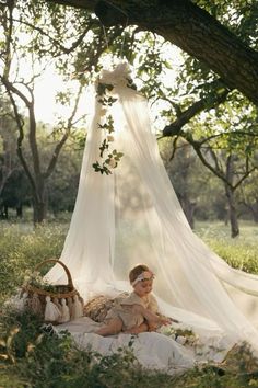 a woman is sitting in the grass under a white canopy with flowers on it and holding a wicker basket