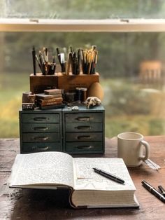 an open book sitting on top of a wooden table next to a cup and pen