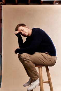 a man sitting on top of a wooden stool in front of a white wall wearing a black sweater and khaki pants