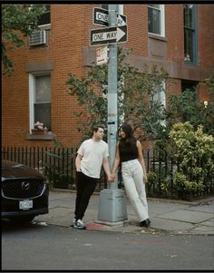 two people standing on the corner of a street holding hands under a one way sign