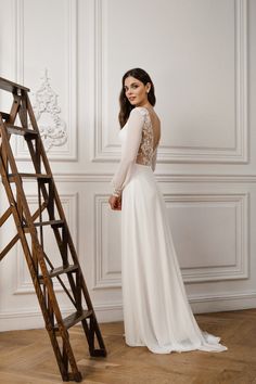 a woman standing next to a ladder wearing a white dress with long sleeves and an open back