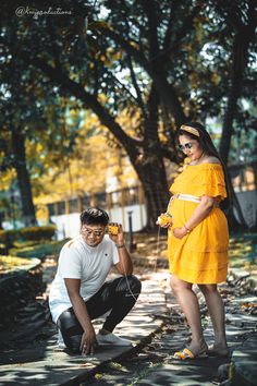 a man kneeling down next to a woman in a yellow dress on the ground near trees