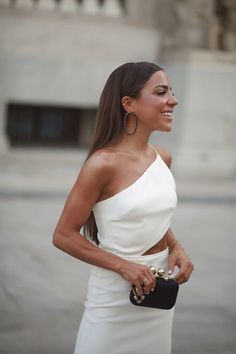 a woman in a white dress is smiling and holding a black purse with her right hand