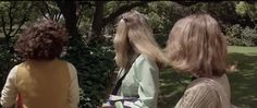 three women standing in front of trees and bushes looking at the ground with one woman's hair blowing back