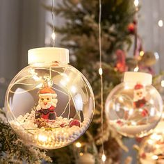 two christmas ornaments hanging from a tree with lights on them and santa claus in a fish bowl