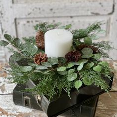 a candle is sitting on top of a black box with greenery and pine cones