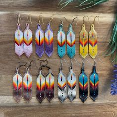 six pairs of beaded earrings on a wooden table next to purple flowers and pine needles