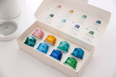 an open box with seven different colored tea cups in it on a white table top