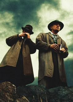 two men standing on top of a rocky hill under a cloudy sky with one holding his coat open