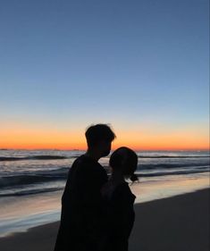 a man and woman standing on top of a beach next to the ocean at sunset