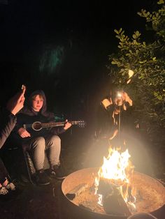 two people sitting around a campfire playing guitar and singing into the fire pit at night