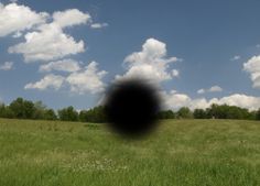 a black object in the middle of a grassy field with trees and clouds behind it