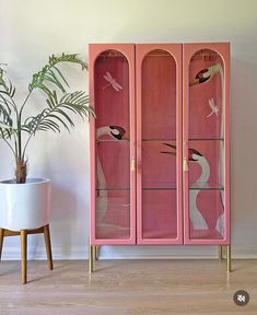 a pink cabinet with birds painted on the glass doors and two potted plants next to it