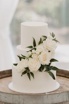 a wedding cake with white flowers and greenery on the top is sitting on a wooden barrel