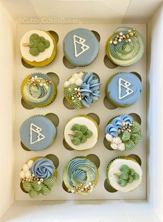 twelve cupcakes in a box decorated with blue and green frosting, including trees