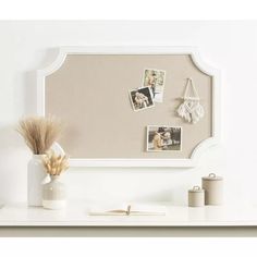 a white desk topped with a vase filled with dried flowers and pictures on the wall