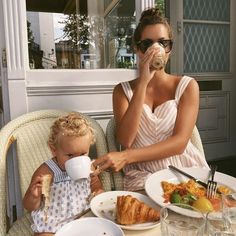 a woman sitting at a table with a child eating food and drinking from a cup