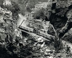 an old black and white photo of a bridge over a stream in the mountains with people on it